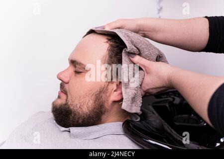 Friseur, der die Haare des Kunden im Friseurladen wäscht. Ein junger bärtiger Mann mit kaukasischem Aussehen an einem Empfang in einem Friseurladen. Der Prozess des Schneidens Stockfoto