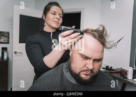 Ein junger bärtiger Mann mit einem Mohawk, der sich vom Friseur auf dem Stuhl im Friseursalon einen Haarschnitt gemacht hat. Ein Friseurmädchen macht einem Typen einen Mohawk. Stockfoto