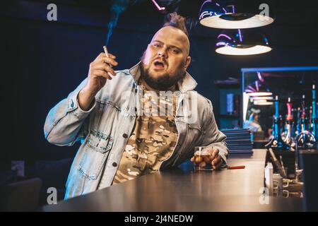 Ein informeller junger Mann mit einem Mohawk auf dem Kopf raucht und trinkt Whiskey in einer Nachtclub-Bar. Schlechte Gewohnheiten. Das Konzept der Alkoholabhängigkeit. Stockfoto