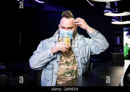 Ein Punk-Mann mit einem Mohawk, der eine Schutzmaske auf seinem Gesicht trägt, trinkt im Dunkeln Bier aus einem Glaskrug. Quarantäne in der Leiste. Alkoholverbot Stockfoto