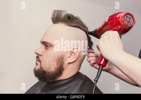 Weibliche Barbier macht eine Mohawk Frisur bei dem erwachsenen Mann mit Bart. Haar-Styling mit einem Haartrockner. Stockfoto