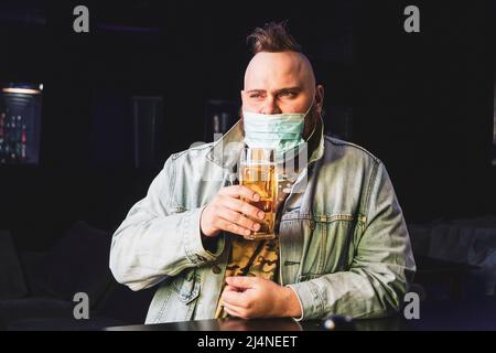 Ein Punk-Mann mit einem Mohawk, der eine Schutzmaske auf seinem Gesicht trägt, trinkt im Dunkeln Bier aus einem Glaskrug. Quarantäne in der Leiste. Alkoholverbot Stockfoto