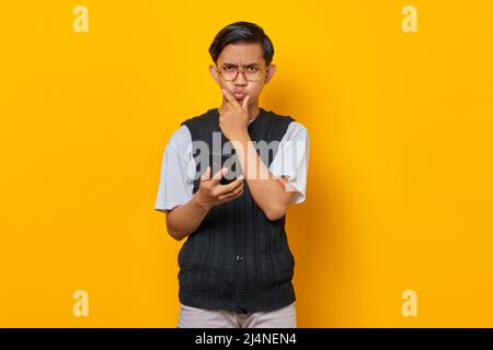 Wütender junger asiatischer Mann mit Finger am Kinn und Mobiltelefon auf gelbem Hintergrund Stockfoto