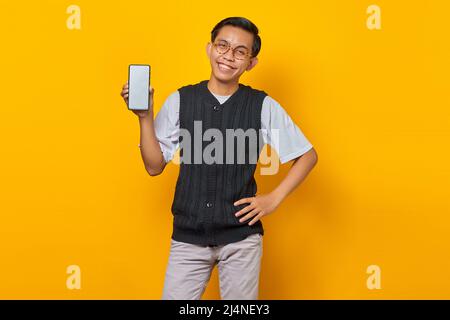 Lächelnder junger, hübscher Mann, der auf gelbem Hintergrund einen leeren Bildschirm des Smartphones in der Hand zeigt Stockfoto