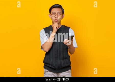 Nachdenklicher junger asiatischer Mann mit Mobiltelefon und Fingern am Kinn auf gelbem Hintergrund Stockfoto