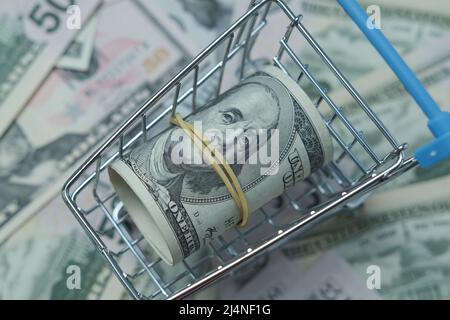 In einen Schlauch gerollte Dollars liegen in einem Einkaufskorb. Der Korb steht auf einem verschwommenen Geldhintergrund. Stockfoto
