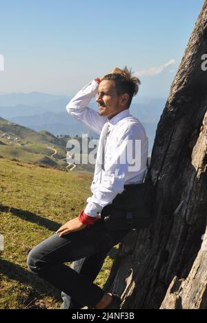 Seitenansicht eines gut aussehenden jungen Mannes, der in ​mountain posiert, seine Hand auf den Haaren bewegt und sich am Baum lehnt Stockfoto