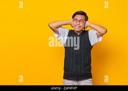 Frustrierter junger Mann aus Asien, der die Ohren mit isolierten Händen auf gelbem Hintergrund bedeckt Stockfoto