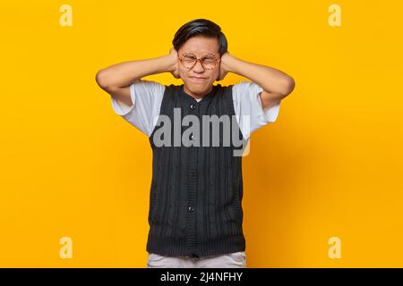 Frustrierter junger Mann aus Asien, der die Ohren mit isolierten Händen auf gelbem Hintergrund bedeckt Stockfoto