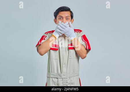 Schockiert junge gutaussehende Mechaniker bedeckt den Mund mit Händen auf grauem Hintergrund Stockfoto