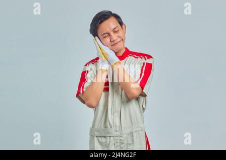 Porträt eines fröhlichen, gutaussehenden Mannes, der mechanische Uniform mit schlafender Geste auf grauem Hintergrund trägt Stockfoto