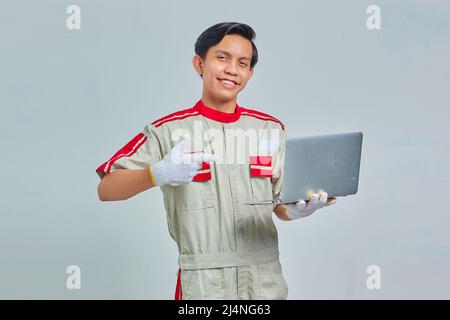 Lächelnder, hübscher junger Mechaniker, der Uniform trägt und auf einen Laptop auf grauem Hintergrund zeigt Stockfoto