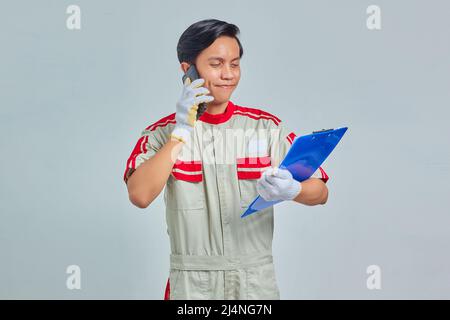 Porträt eines lächelnden jungen asiatischen Mechanikers, der am Telefon spricht und Clipboard auf grauem Hintergrund isoliert hält Stockfoto