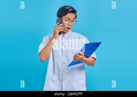 Porträt einer überraschten Krankenschwester, die auf dem Smartphone spricht, während Clipboard auf blauem Hintergrund isoliert gehalten wird Stockfoto