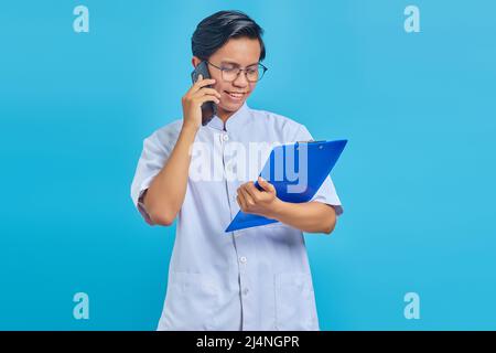 Portrait einer Krankenschwester, die lächelt und auf dem Smartphone spricht, während sie Clipboard auf blauem Hintergrund isoliert hält Stockfoto