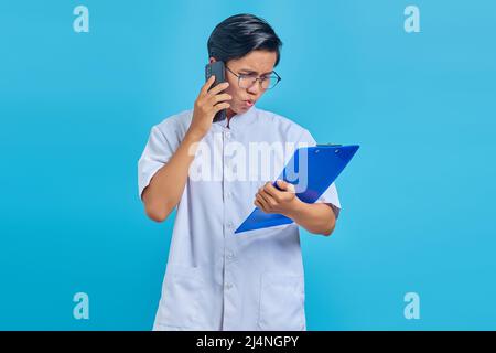 Portrait einer Krankenschwester, die lächelt und auf dem Smartphone spricht, während sie Clipboard auf blauem Hintergrund isoliert hält Stockfoto