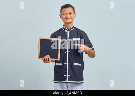 Porträt eines fröhlichen asiatischen jungen Mannes in Karate-Uniform, der auf ein leeres Brett mit Finger auf grauem Hintergrund zeigt Stockfoto