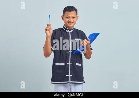 Porträt eines lächelnden asiatischen jungen Mannes, der Karate-Uniform trägt, die Zwischenablage hält und mit einem Stift nach oben zeigt Stockfoto