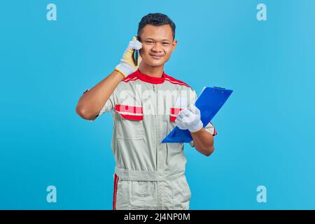 Lächelnder junger asiatischer Mechaniker, der auf dem Handy spricht und Clipboard auf blauem Hintergrund isoliert hält Stockfoto