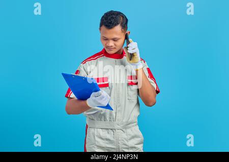 Ernst aussehende junge asiatische Mechaniker reden auf Handy und Überprüfung von Notizen auf Zwischenablage isoliert auf blauem Hintergrund Stockfoto