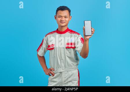 Porträt eines lächelnden jungen Mechanikers, der einen leeren Smartphone-Bildschirm in der Hand zeigt, isoliert auf blauem Hintergrund Stockfoto