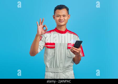 Porträt eines lächelnden jungen Mechanikers, der eine Geste in Ordnung gebracht hat und das auf blauem Hintergrund isolierte Genehmigungssymbol zeigt Stockfoto