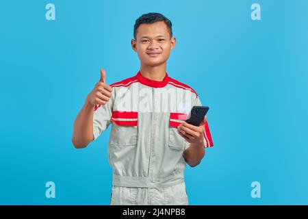 Porträt eines lächelnden jungen Mechanikers, der auf blauem Hintergrund eine Geste mit dem Daumen zeigt Stockfoto