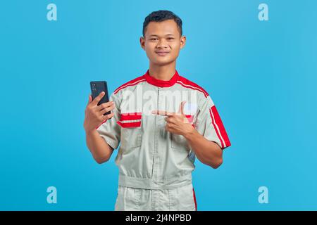 Porträt eines lächelnden jungen Mechanikers, der mit dem Finger auf das auf blauem Hintergrund isolierte Mobiltelefon zeigt Stockfoto