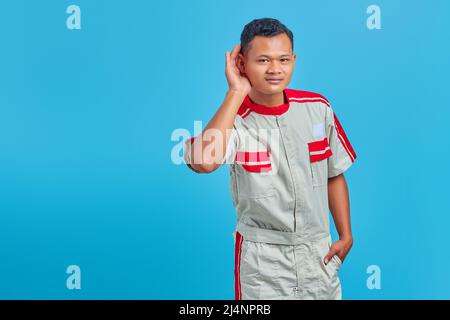 Ernst aussehende asiatische Mechaniker versuchen, geheime Unterhaltung isoliert auf blauem Hintergrund zu hören Stockfoto