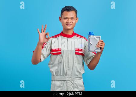 Porträt eines lächelnden jungen asiatischen Mechanikers, der eine Plastikflasche Motoröl hält und mit dem Daumen nach oben auf blauem Hintergrund die Genehmigung zeigt Stockfoto