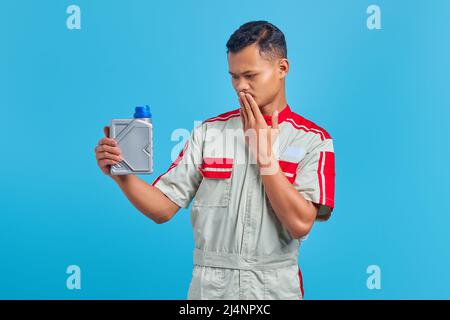 Schockiert junge asiatische Mechaniker Porträt hält Motoröl Plastikflasche und deckt Mund auf blauen Hintergrund Hände Stockfoto