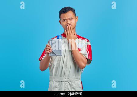 Schockiert junge asiatische Mechaniker Porträt hält Motoröl Plastikflasche und deckt Mund auf blauen Hintergrund Hände Stockfoto