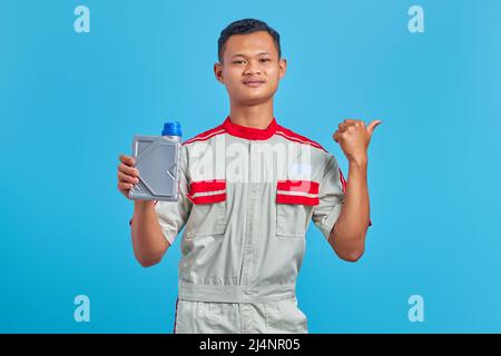 Porträt eines lächelnden jungen asiatischen Mechanikers, der eine Plastikflasche mit Motoröl zeigt und auf leeren Raum auf blauem Hintergrund zeigt Stockfoto