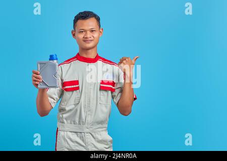 Porträt eines lächelnden jungen asiatischen Mechanikers, der eine Plastikflasche mit Motoröl zeigt und auf leeren Raum auf blauem Hintergrund zeigt Stockfoto