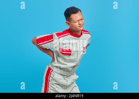 Porträt eines jungen asiatischen Mechanikers, der unter Rückenschmerzen leidet und auf blauen Händen auf dem Hintergrund den Rücken berührt Stockfoto