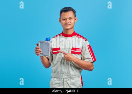 Porträt eines lächelnden jungen asiatischen Mechanikers, der auf eine Plastikflasche Motoröl zeigt, mit dem Finger auf blauem Hintergrund Stockfoto