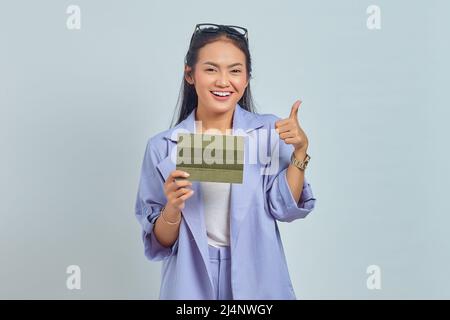 Porträt einer fröhlichen jungen asiatischen Frau, die Fahrzeugbuch hält und die Daumen nach oben zeigt, Geste isoliert auf weißem Hintergrund Stockfoto