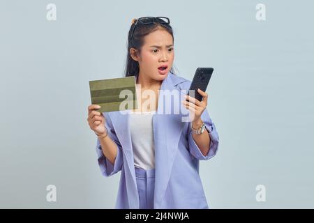Porträt einer überraschten jungen asiatischen Frau, die Fahrzeugbuch zeigt und Smartphone isoliert auf weißem Hintergrund hält Stockfoto