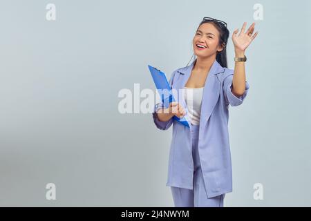 Porträt einer fröhlichen jungen asiatischen Geschäftsfrau, die einen Dokumentenordner in der Hand hält, mit ihren Handflächen winkt und Freunde begrüßt und ein freundliches Schild auf pur zeigt Stockfoto