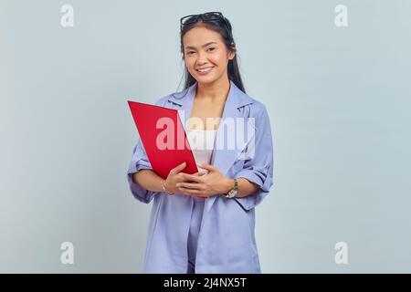 Porträt einer jungen asiatischen Geschäftsfrau, die selbstbewusst mit Ordnerdokumenten steht und lächelnd auf die Kamera blickt, die auf violettem Hintergrund isoliert ist Stockfoto