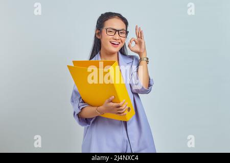 Porträt einer fröhlichen jungen asiatischen Geschäftsfrau, die Dokumentenmappe hält und auf purpurem Hintergrund eine Geste zeigt, die in Ordnung ist Stockfoto