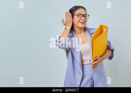 Porträt einer ernsthaften jungen asiatischen Geschäftsfrau hält die Hand in der Nähe des Ohrs versucht, private Gespräche zu hören, lauscht Klatsch und hält einen gelben Ordner Stockfoto