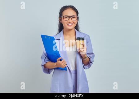 Porträt einer fröhlichen jungen asiatischen Geschäftsfrau, die Zwischenablage hält und Kaffee zum Mitnehmen trinkt, isoliert auf violettem Hintergrund Stockfoto