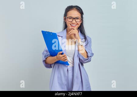 Porträt einer fröhlichen jungen asiatischen Geschäftsfrau, die das Kinn hält, während sie den Dokumentenordner auf violettem Hintergrund isoliert hält Stockfoto