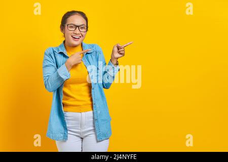 Porträt einer fröhlichen jungen asiatischen Studentin in Denim-Kleidung, die mit dem Finger auf den Kopierraum zeigt und die Kamera auf gelbem Hintergrund isoliert betrachtet Stockfoto