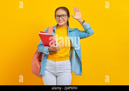 Lächelnde junge asiatische Studentin in Denim-Kleidung mit Rucksack, die Notizbuch hält, winkt und grüßt mit der Hand, während jemand auf Gelb isoliert bemerkt Stockfoto