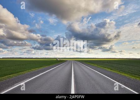 Leere Asphaltstraße außerhalb der Stadt zwischen grünen Feldern und einem schönen bewölkten Himmel Stockfoto