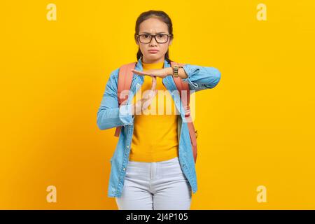 Schöne junge asiatische Frau Student in Denim-Outfit mit Rucksack zeigt Auszeit Geste mit Händen isoliert auf gelbem Hintergrund. Ausbildung in Univ Stockfoto