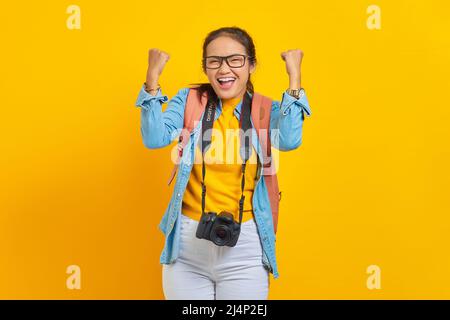 Porträt der aufgeregten jungen Reisenden asiatischen Frau mit Rucksack und Kamera in Denim-Kleidung, während feiern Glück isoliert auf gelbem Hintergrund. Passagier Stockfoto