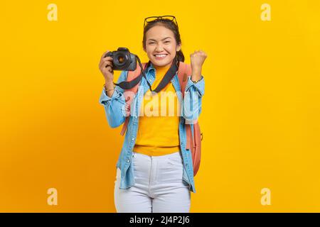 Porträt einer aufgeregten jungen asiatischen Frau mit Rucksack, der eine professionelle Kamera hält und auf gelbem Hintergrund das Glück feiert Stockfoto